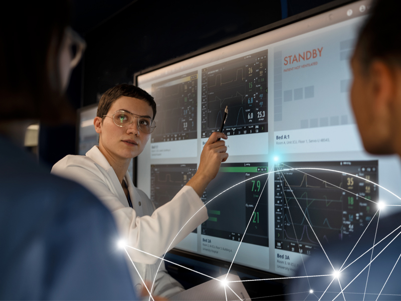 Person in a lab coat points at a screen displaying technical graphs and data, with others in discussion.