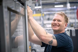 Lächelnder Mann mit Tätowierungen, der in einem industriellen Umfeld an Maschinen arbeitet.