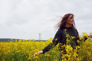 A woman with curly hair stands in a field of yellow flowers, wearing a black top, with cloudy sky in the background.
