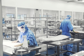 Scientists in blue protective suits work in a cleanroom with stainless steel tables and shelves.