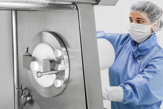 Person in protective gear operates a large stainless steel machine in a sterile laboratory environment.