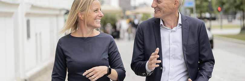 Zwei Menschen gehen und unterhalten sich auf einer Straße, beide lächeln und tragen Geschäftskleidung.