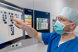 Ein Chirurg im OP-Kittel und mit Maske bedient einen Touchscreen in einer medizinischen Einrichtung mit mehreren Monitoren.