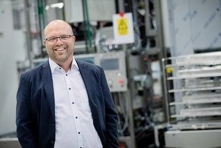 Lächelnder Mann in Anzug und Brille, der in einer Industrieanlage mit Maschinen im Hintergrund steht.