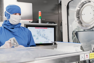 Een persoon in blauwe beschermende kleding bedient machines in een steriele laboratoriumomgeving, met een monitor op de achtergrond.