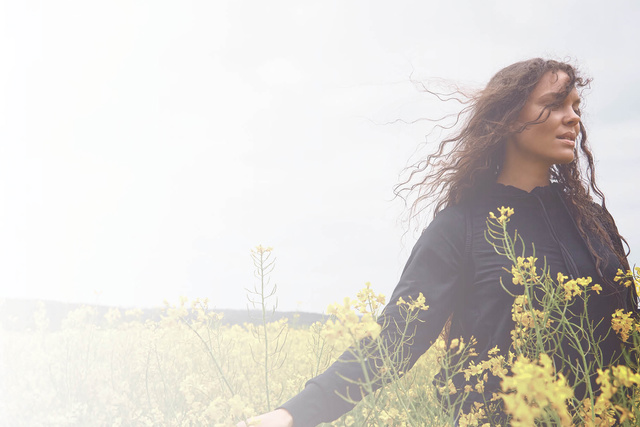 A woman with curly hair is in a field of yellow flowers. She is wearing a black top, and there is a cloudy sky in the background