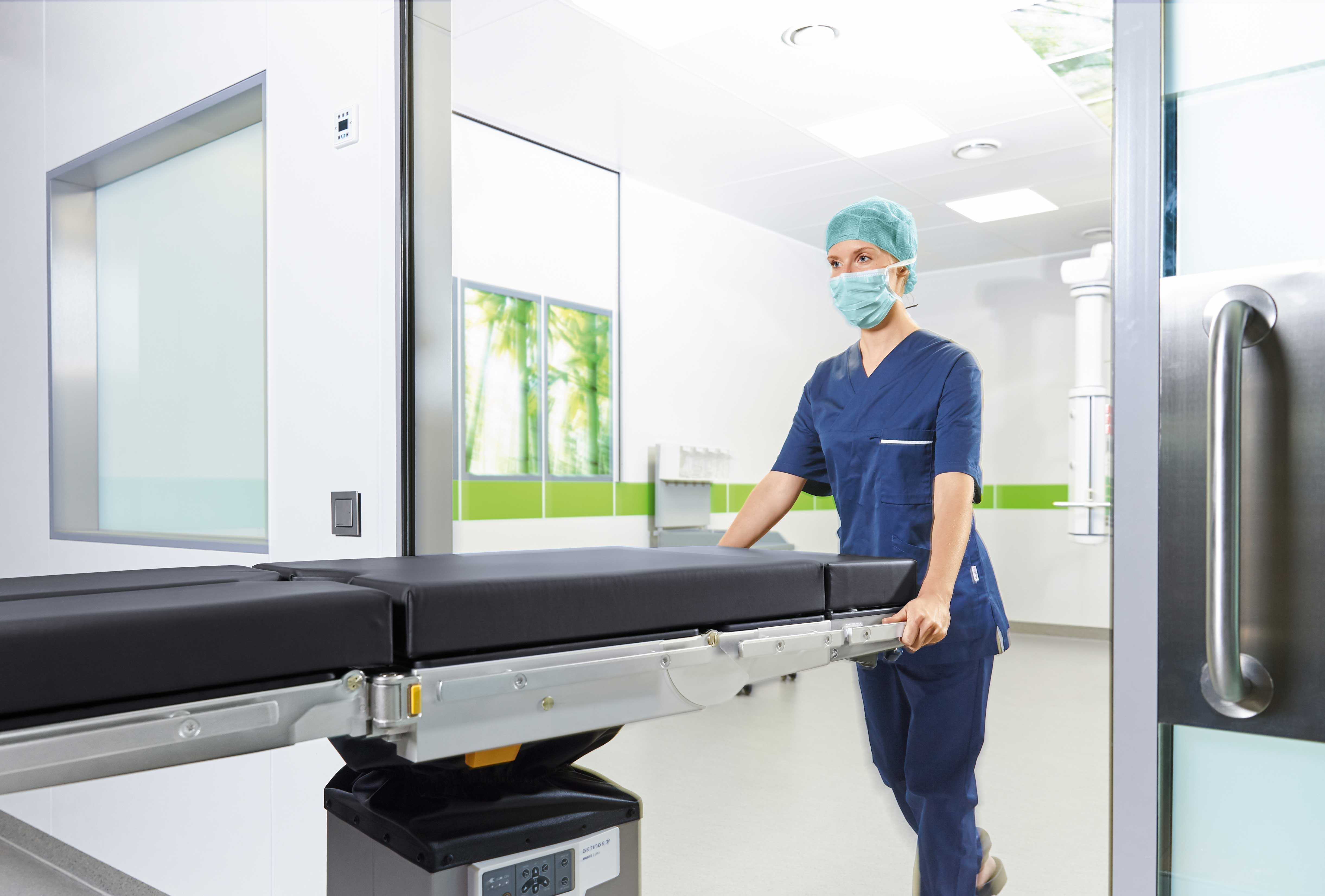 Healthcare worker in scrubs and mask pushing a hospital bed in a bright, modern medical facility.