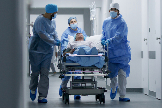 A team of healthcare workers in blue scrubs and masks are pushing a hospital bed along a hallway in a medical facility.