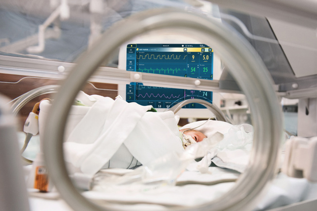 Neonate in incubator with Getinge Servo-n mechanical ventilator screen seen in the background