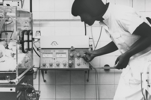 A clinician adjusting the pause time setting on a Servo 900 mechanical ventilator placed next to an a baby in an incubator 