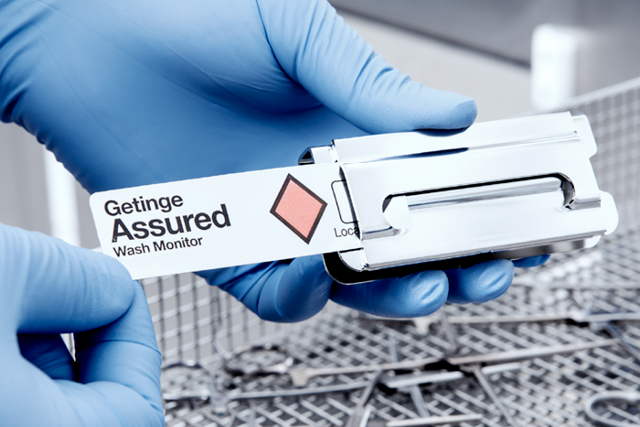 Gloved hands hold a Getinge Assured Wash Monitor over medical instruments in a sterilization setting.