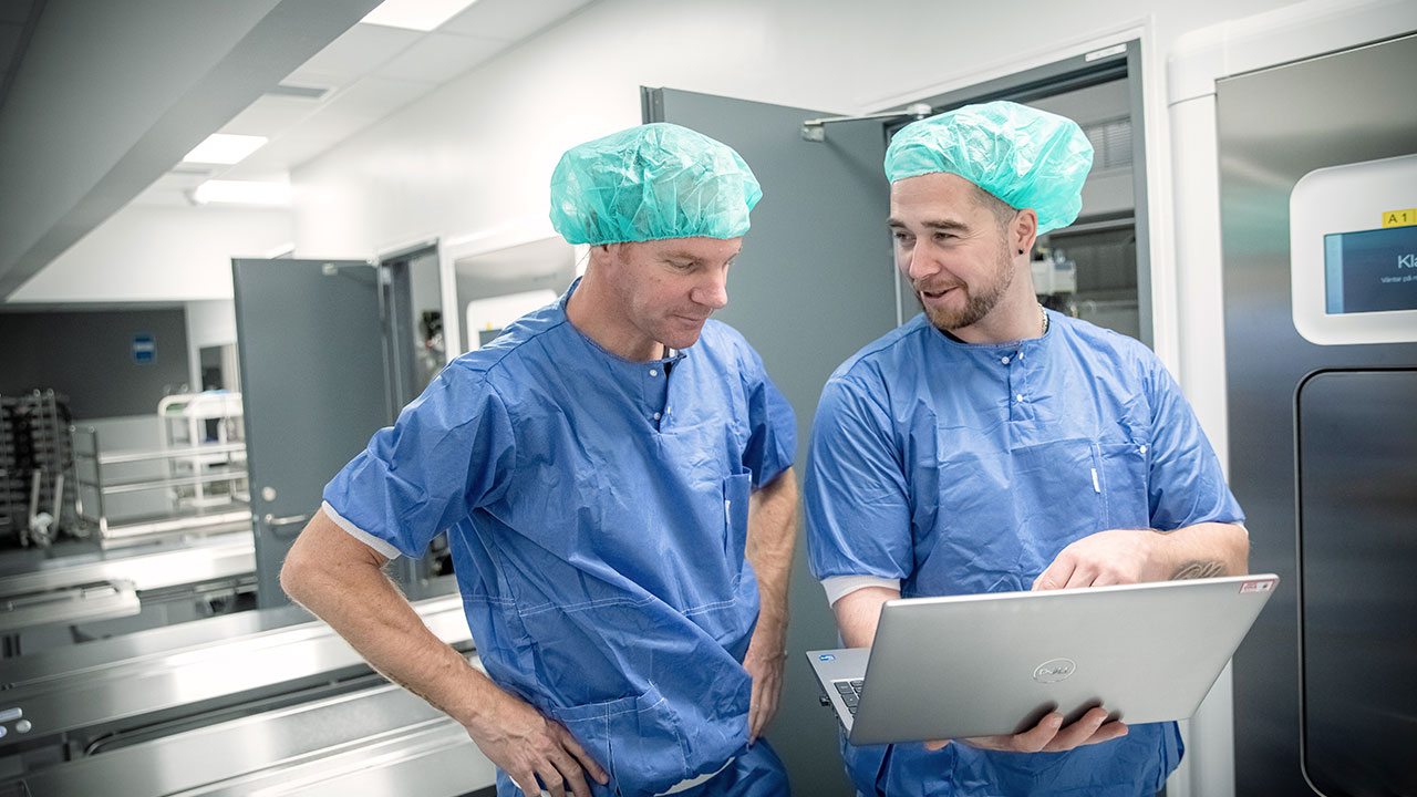 Two surgeons in surgical scrubs focused on a laptop screen in a clinical environment.