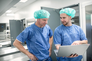 Two surgeons in surgical scrubs focused on a laptop screen in a clinical environment.